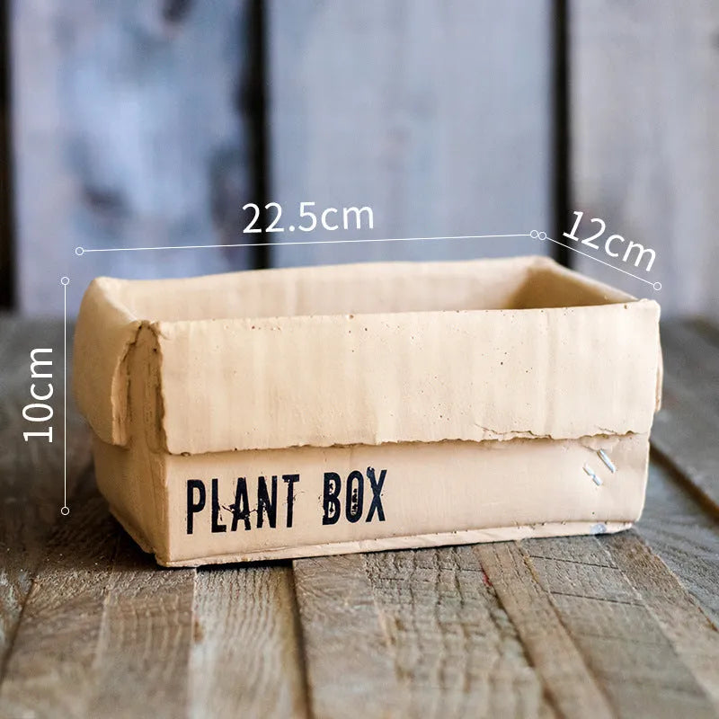 A grey cement flowerpot with a folded paper box design, shown with a drainage hole in its base.