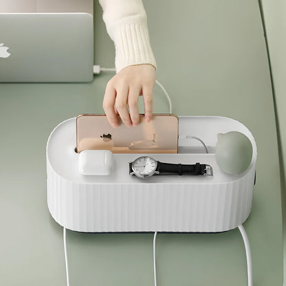 Blue desktop cable storage box with a white lid, showing cable ports on the base and a mobile phone sitting in its slot on top.