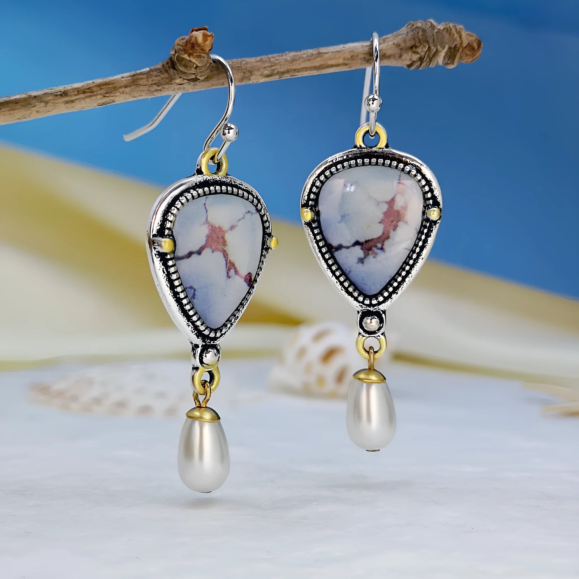 Close-up of retro blue geometric drop earrings with a synthetic blue opal stone and white pearl accent on an electroplated metal frame.