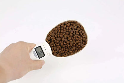 White electronic pet feeding weighing spoon with digital display and removable metal shovel head on a table.