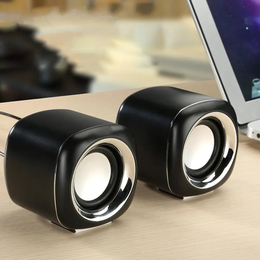 A pair of small black or white cube-shaped wired computer speakers with a 3.5mm audio plug on a desk.