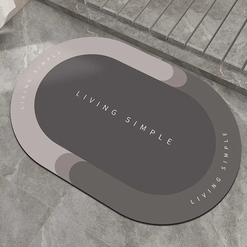Oval grey bath mat with non-slip rubber bottom on a tiled floor, showing absorbent fabric surface