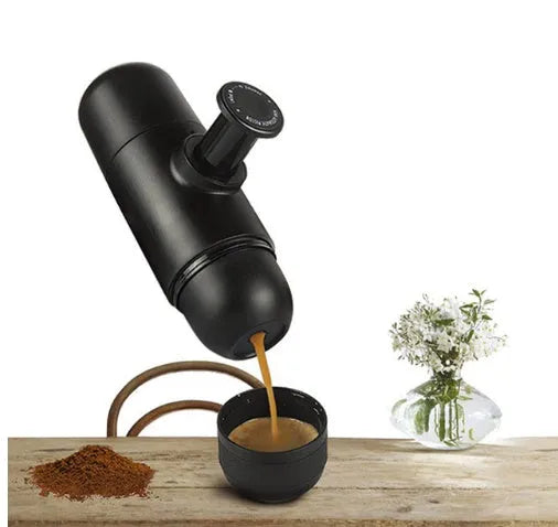 Black portable manual espresso maker with stainless steel parts, shown next to a coffee bag and mug.