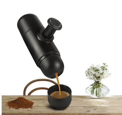 Black portable manual espresso maker with stainless steel parts, shown next to a coffee bag and mug.