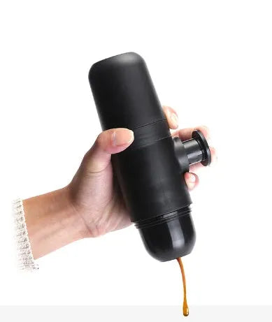 Black portable manual espresso maker with stainless steel parts, shown next to a coffee bag and mug.