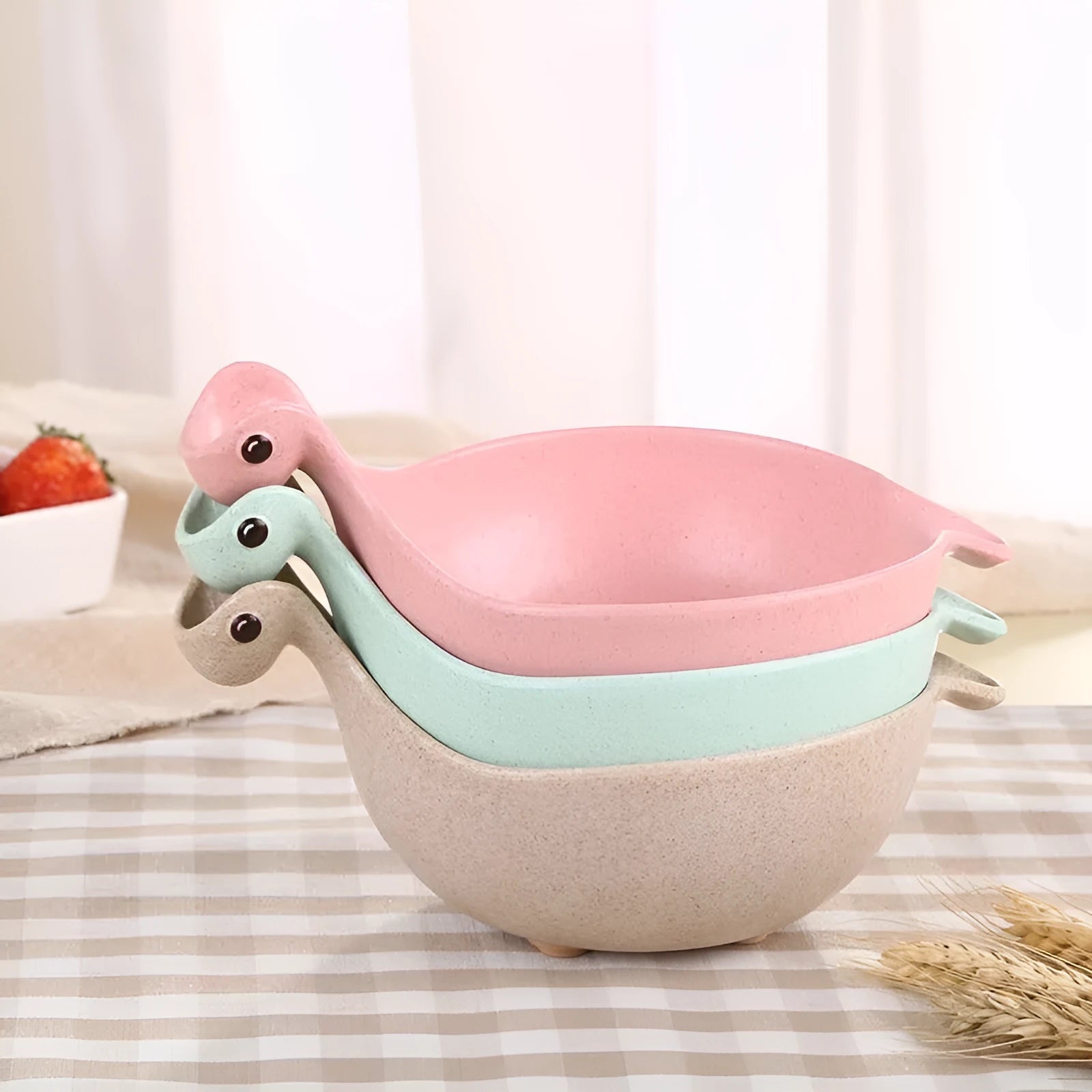 Three colourful dinosaur-shaped bowls made from wheat straw in pink, green, and beige.