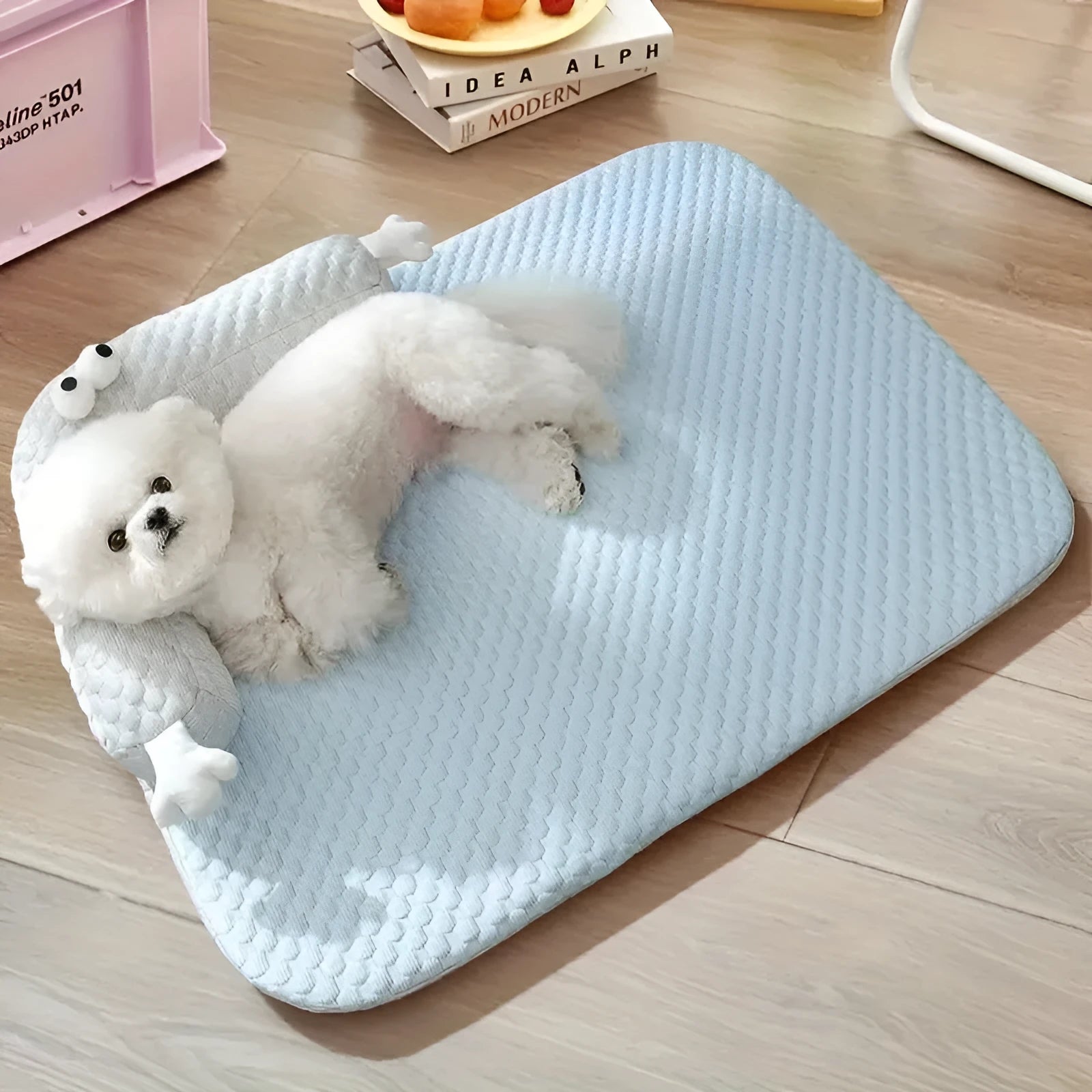 Blue pet bed cushion with big-eyed monster design, shown on a wooden floor.