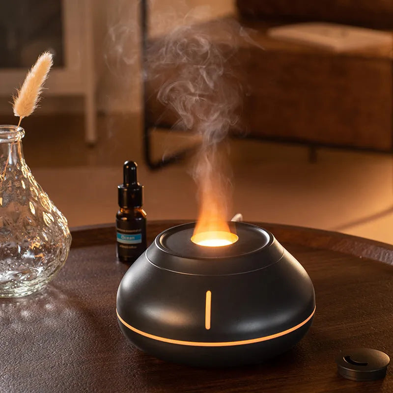 Round white ultrasonic aroma diffuser with colourful LED flame effect and mist emitting from the top on a wooden desk.