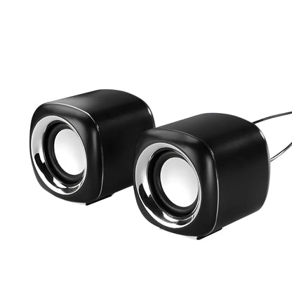 A pair of small black or white cube-shaped wired computer speakers with a 3.5mm audio plug on a desk.