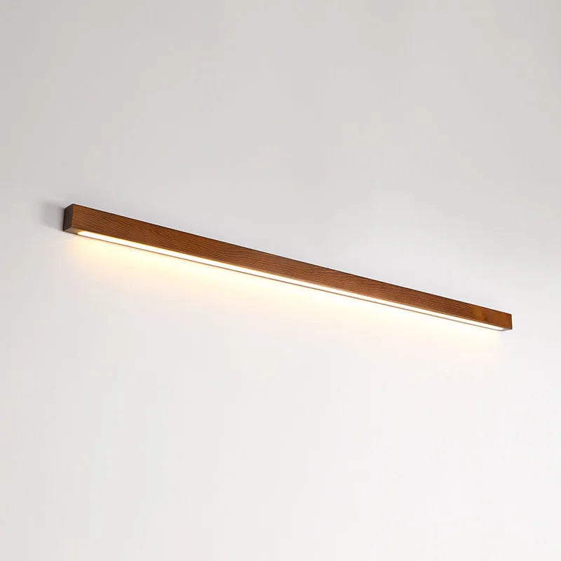 Slim walnut-coloured wooden strip wall lamp with acrylic light diffuser mounted on a plain wall.
