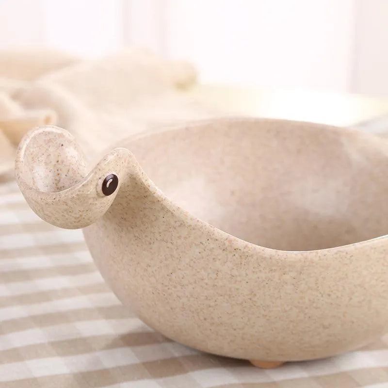 Three colourful dinosaur-shaped bowls made from wheat straw in pink, green, and beige.
