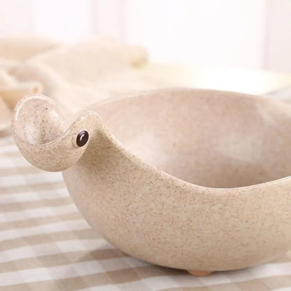 Three colourful dinosaur-shaped bowls made from wheat straw in pink, green, and beige.
