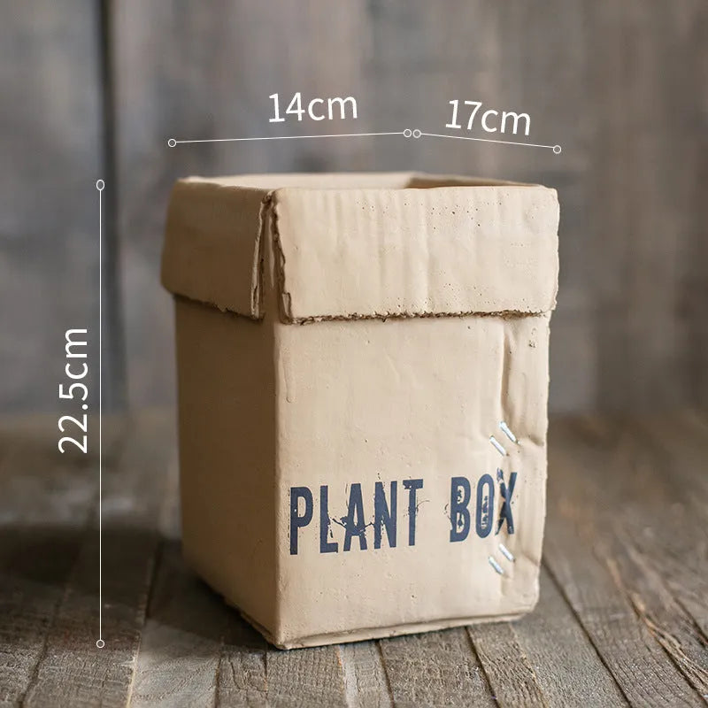 A grey cement flowerpot with a folded paper box design, shown with a drainage hole in its base.