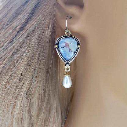 Close-up of retro blue geometric drop earrings with a synthetic blue opal stone and white pearl accent on an electroplated metal frame.