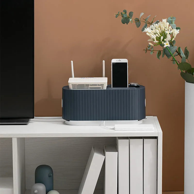 Blue desktop cable storage box with a white lid, showing cable ports on the base and a mobile phone sitting in its slot on top.