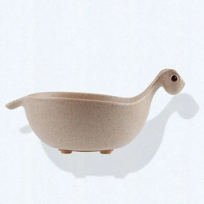 Three colourful dinosaur-shaped bowls made from wheat straw in pink, green, and beige.