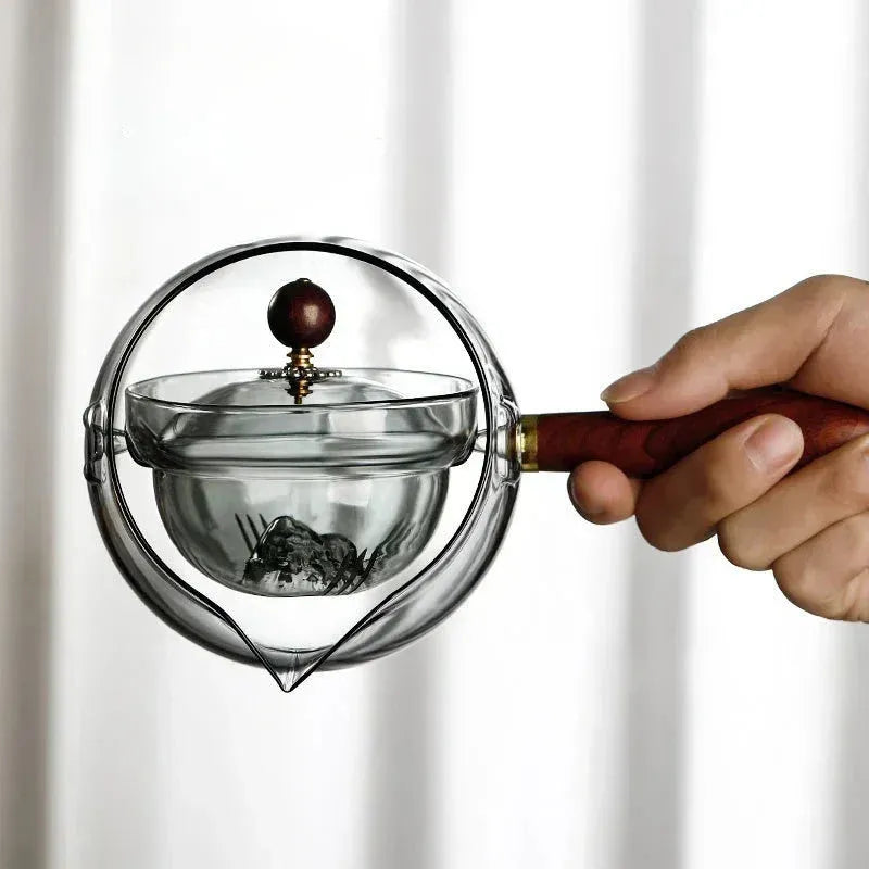 Transparent borosilicate glass rotary teapot with wooden handle and built-in infuser on a wooden surface