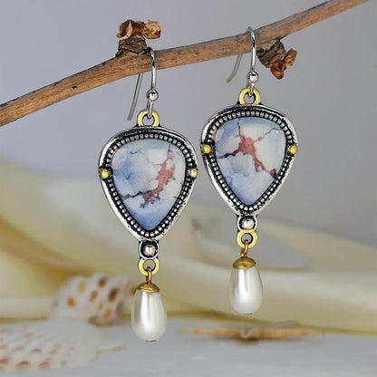 Close-up of retro blue geometric drop earrings with a synthetic blue opal stone and white pearl accent on an electroplated metal frame.