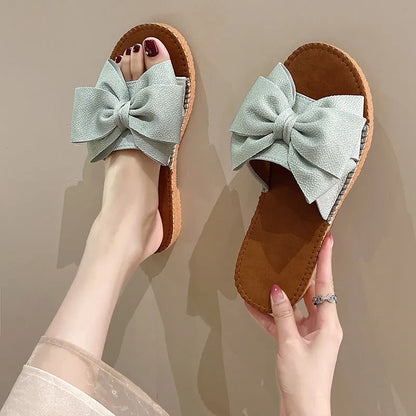 Women's beige suede slip-on sandals with a bow detail and soft rubber flat sole.