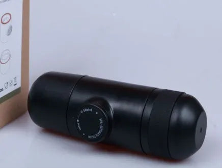 Black portable manual espresso maker with stainless steel parts, shown next to a coffee bag and mug.