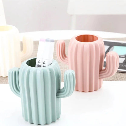 A white cactus-shaped desktop organiser holding pens, pencils, and makeup brushes on a wooden desk